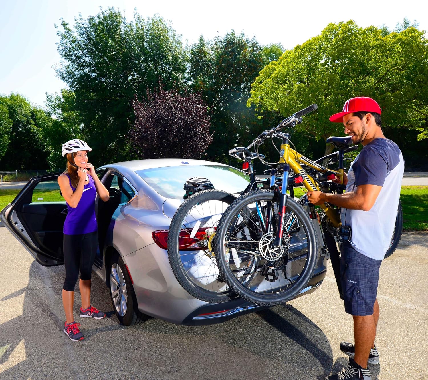 Two people attaching bicycles to a rear trunk-mounted bike rack on a car for outdoor cycling transport.
